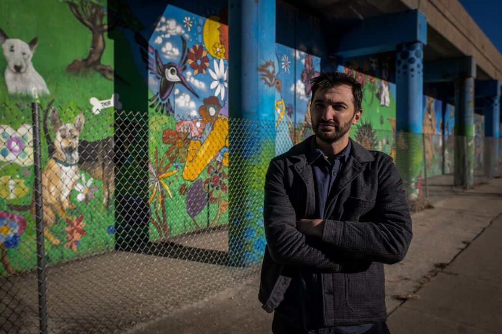 A man with stands with his arms crossed in front of a colorful mural where there's dogs, hummingbirds, butterflies, and flowers.
