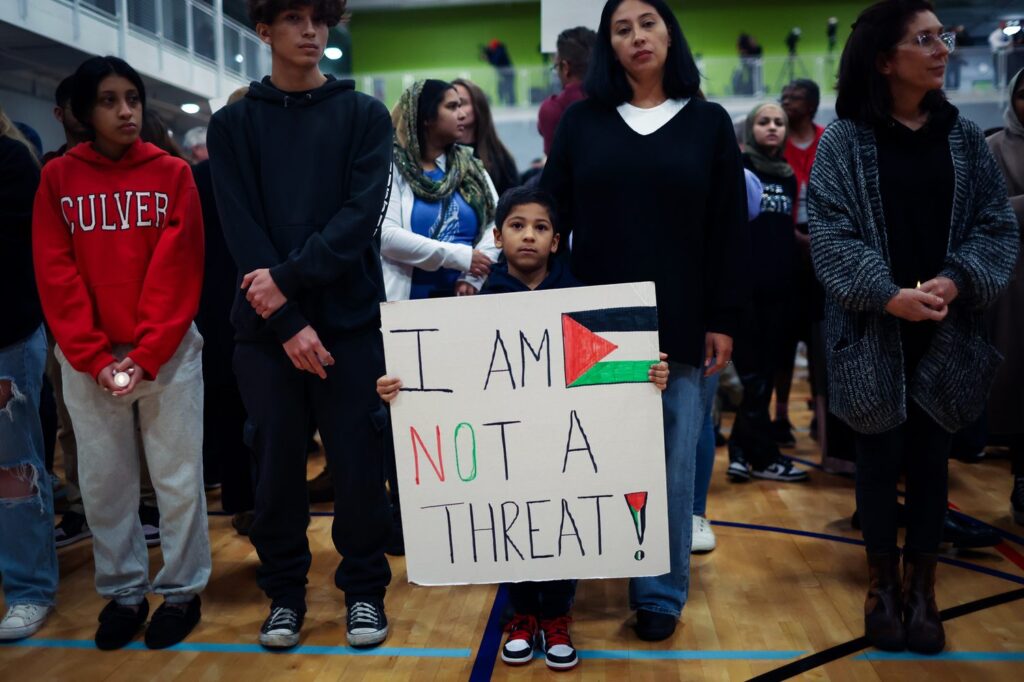 A group of people stand on a wooden floor. In the middle, a small boy is holding a banner with the Palestinian flag that reads,