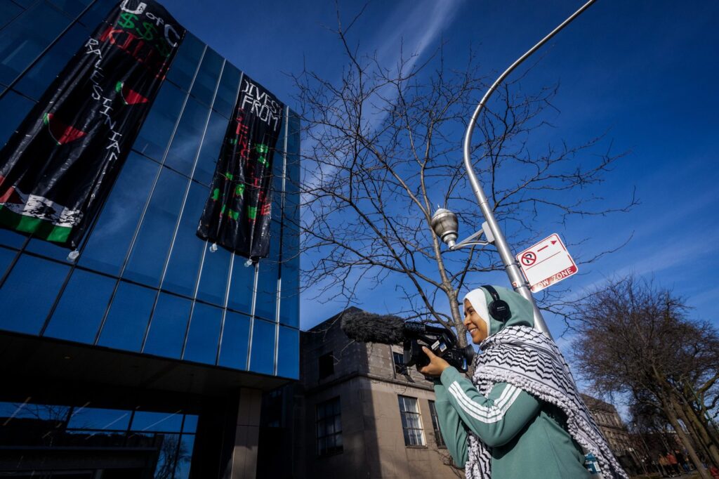 A woman wearing headphones holds a camera, filming a building with banners that read,